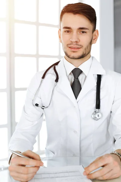 Médecin homme remplir les dossiers de l'histoire des médicaments formulaire alors qu'il était assis au bureau en verre dans une clinique ensoleillée. Concept de médecine — Photo