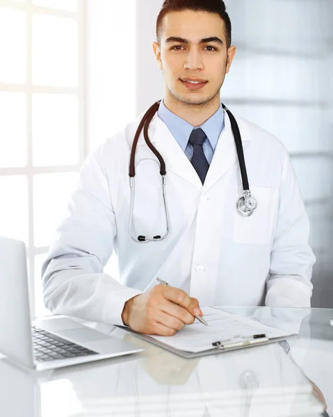 Homem médico árabe está usando computador portátil ao preencher registros de histórico de medicação forma clínica ensolarada. Conceito de medicina — Fotografia de Stock