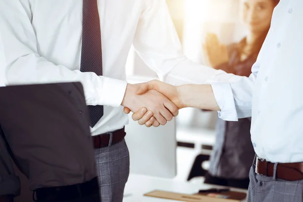 Two businessmen shaking hands in sunny office, close-up. Happy and excited business woman stands with raising hands at the background. Business people concept — Stock Photo, Image