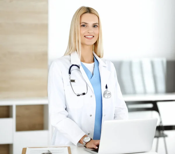 금발의 여성 의사가 랩탑 컴퓨터를 사용하면서 웃고 있다. 병원에서 일하는 여성 - 의사들은 그녀의 직업에 대해 흥분하고 행복해 했습니다. 의학적 개념 — 스톡 사진