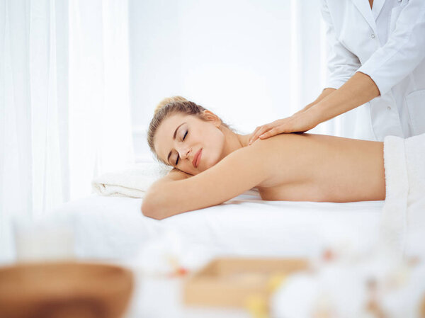 Young and blonde woman enjoying massage of back in spa salon. Beauty concept