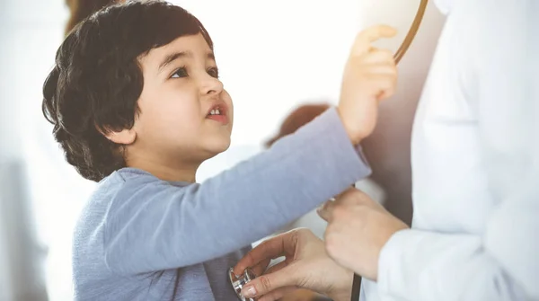 Dokter-vrouw die een kindpatiënt onderzoekt met stethoscoop in een zonnige kliniek. Leuke arabische jongen en zijn broer op afspraak arts — Stockfoto