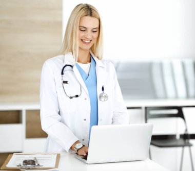 Sarışın kadın doktor dizüstü bilgisayar kullanırken gülümsüyor. Klinikteki kadın doktor mesleğinden dolayı heyecanlı ve mutlu. Tıp konsepti