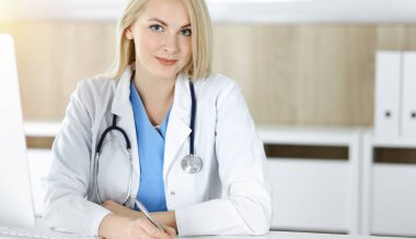 Güneşli klinikte masa başında oturan kadın doktor. Sarışın, neşeli doktor ilaç geçmişi formunu dolduruyor.