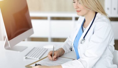 Güneşli klinikte masa başında oturan kadın doktor. Sarışın, neşeli doktor ilaç geçmişi formunu dolduruyor.