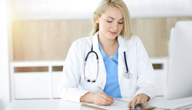 Güneşli klinikte masa başında oturan kadın doktor. Sarışın, neşeli doktor ilaç geçmişi formunu dolduruyor.
