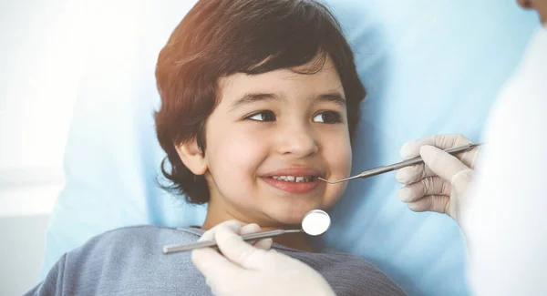 Lindo chico árabe sentado en la silla dental con la boca abierta durante el control oral con el médico. Visita al soleado consultorio del dentista — Foto de Stock