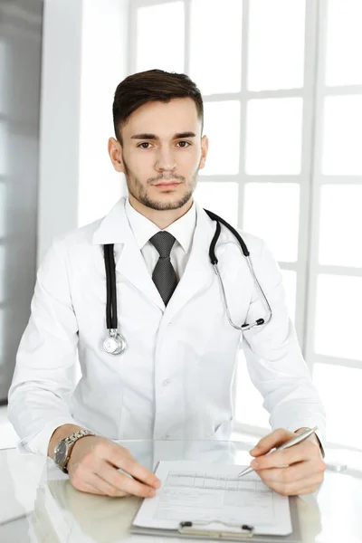 Doktor vyplňuje záznamy o historii léků, zatímco sedí u skleněného stolu na klinice. Medicína a koncept zdravotní péče — Stock fotografie