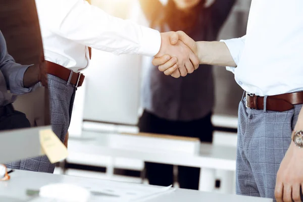 Two businessmen shaking hands in sunny office, close-up. Happy and excited business woman stands with raising hands at the background. Business people concept — Stock Photo, Image