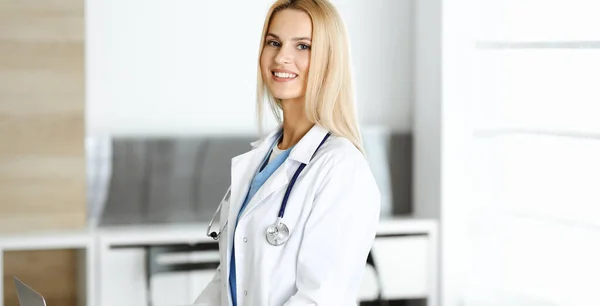 Blond kvinnlig läkare är lsmiling när du använder bärbar dator. Kvinna-läkare på jobbet på kliniken upphetsad och glad över sitt yrke. Medicinskt koncept — Stockfoto