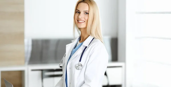 La médica rubia está sonriendo mientras usa la computadora portátil. Mujer-médico en el trabajo en la clínica emocionada y feliz de su profesión. Concepto de medicina — Foto de Stock