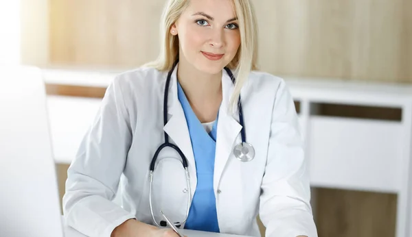 Жінка-лікар на роботі, сидячи за столом у сонячній клініці. Блондинка веселий лікар готовий допомогти — стокове фото