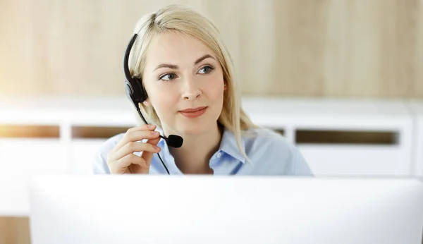 Donna d'affari bionda seduta e comunicata da auricolare in call center in ufficio soleggiato. Concetto di attività di televendita — Foto Stock
