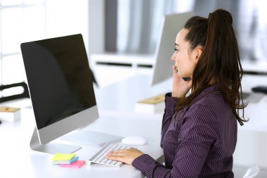 Modern ofisteki iş yerinde telefon ve bilgisayar kullanan bir iş kadını. Esmer sekreter ya da kadın avukat gülümsüyor ve mutlu görünüyor. Zevk ve başarı için çalışmak