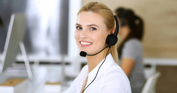 Mujer rubia de negocios con auriculares para la comunicación y la consulta de las personas en la oficina de servicio al cliente. Centro de llamadas. Grupo de operadores en el trabajo en segundo plano — Foto de Stock