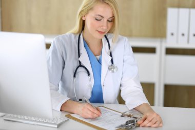 Hastanedeki ya da klinikteki masada oturan doktor kadın. Sarışın, neşeli doktor tıbbi kayıtları dolduruyor. Veriler ve tıp ve sağlık hizmetlerindeki en iyi hizmet