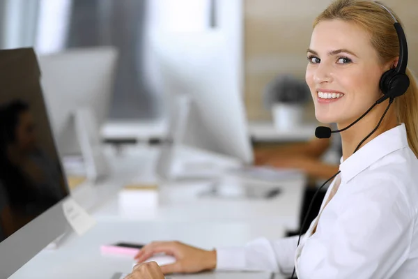 金发女商人使用耳机与客户服务办公室的人进行交流和咨询。呼叫中心背景下工作的操作员小组 — 图库照片