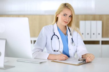 Hastanedeki ya da klinikteki masada oturan doktor kadın. Sarışın, neşeli doktor tıbbi kayıtları dolduruyor. Veriler ve tıp ve sağlık hizmetlerindeki en iyi hizmet