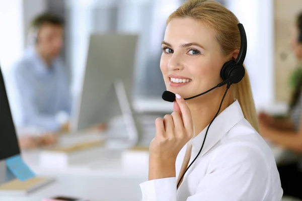 Mujer rubia de negocios con auriculares para la comunicación y la consulta de las personas en la oficina de servicio al cliente. Centro de llamadas. Grupo de operadores en el trabajo en segundo plano —  Fotos de Stock