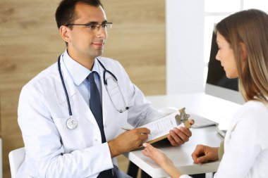 Doktor ve hasta klinikteki bir masada otururken fiziksel muayenenin sonuçlarını tartışıyorlar. Genç bir kadının tıbbi geçmişini doldurmak için pano kullanan bir erkek doktor.