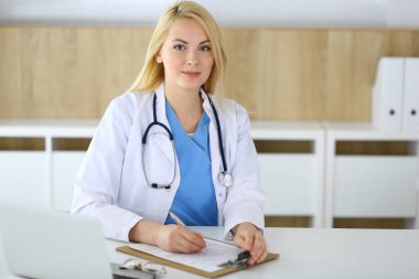 Hastanedeki ya da klinikteki masada oturan doktor kadın. Sarışın, neşeli doktor tıbbi kayıtları dolduruyor. Veriler ve tıp ve sağlık hizmetlerindeki en iyi hizmet