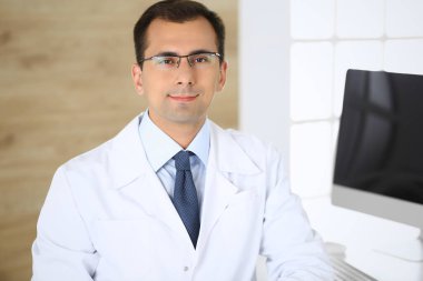 Doktor adam çalışma yerinde masada oturuyor ve kameraya gülümsüyor. Klinikte mükemmel bir tıbbi hizmet. Tıbbın ve sağlık hizmetlerinin mutlu geleceği