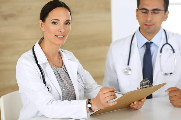 Médico de mediana edad llenando documentos médicos o prescripción con su colega varón. Grupo de médicos en el trabajo discutiendo problemas de tratamiento mientras están sentados en un escritorio en el hospital. Datos en — Foto de Stock