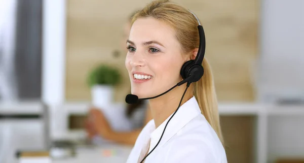 Blondynka bizneswoman za pomocą zestawu słuchawkowego do komunikacji i konsultacji z ludźmi w biurze obsługi klienta. Zadzwoń do centrum. Grupa operatorów w miejscu pracy w tle — Zdjęcie stockowe