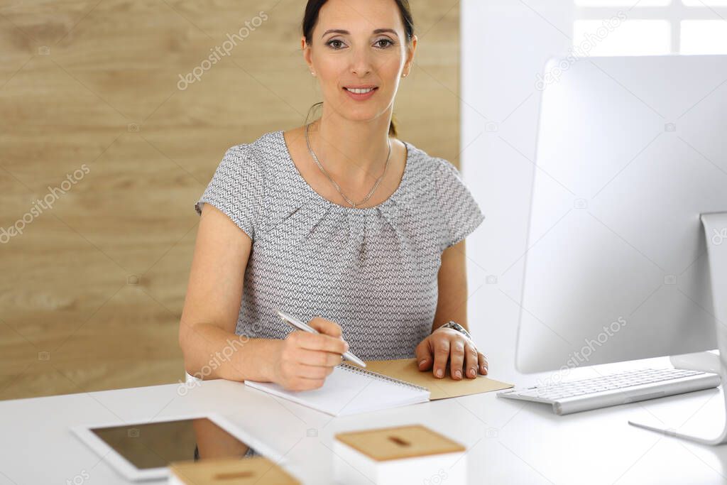 Cheerful smiling business woman working with pc computer while sitting at the desk in modern office. Middle aged female lawyer or auditor at work. Business people concept