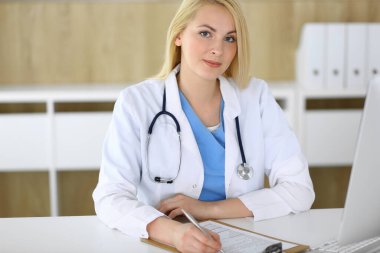 Hastanedeki ya da klinikteki masada oturan doktor kadın. Sarışın, neşeli doktor tıbbi kayıtları dolduruyor. Veriler ve tıp ve sağlık hizmetlerindeki en iyi hizmet