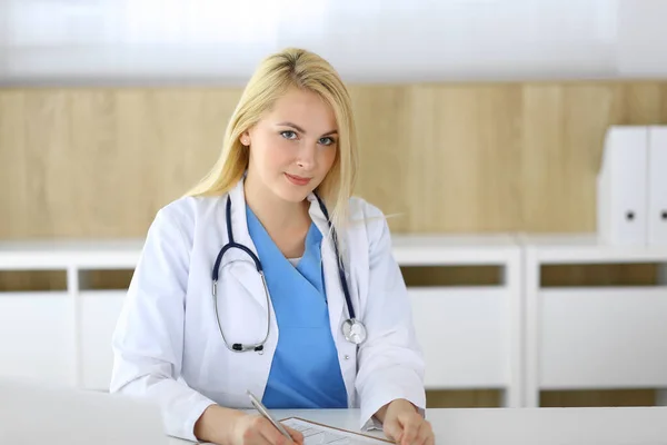 Женщина-врач на работе, сидя за столом в больнице или клинике. Блондинка веселый врач заполняя медицинскую карточку. Данные и лучший сервис в медицине и здравоохранении — стоковое фото