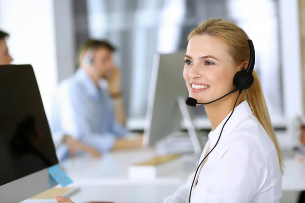 Mujer rubia de negocios con auriculares para la comunicación y la consulta de las personas en la oficina de servicio al cliente. Centro de llamadas. Grupo de operadores en el trabajo en segundo plano — Foto de Stock