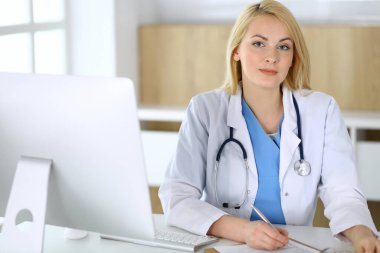 Hastanedeki ya da klinikteki masada oturan doktor kadın. Sarışın, neşeli doktor tıbbi kayıtları dolduruyor. Veriler ve tıp ve sağlık hizmetlerindeki en iyi hizmet