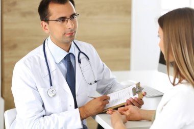 Doktor ve hasta klinikteki bir masada otururken fiziksel muayenenin sonuçlarını tartışıyorlar. Genç bir kadının tıbbi geçmişini doldurmak için pano kullanan bir erkek doktor.