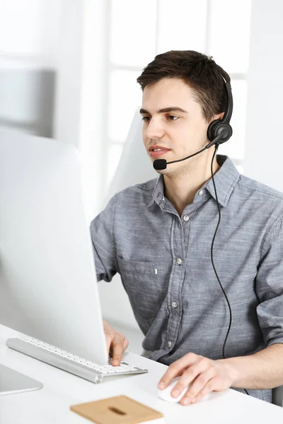 Nenuceně oblečený mladý muž pomocí sluchátka a počítače při rozhovoru se zákazníky on-line. Call centrum, obchodní koncept — Stock fotografie