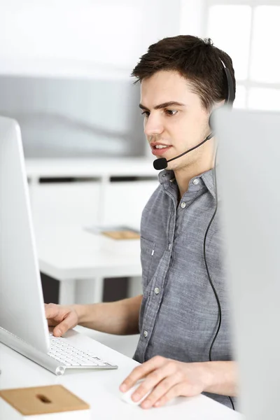 Hombre joven vestido casual usando auriculares y computadora mientras habla con los clientes en línea. Call center, concepto de negocio —  Fotos de Stock