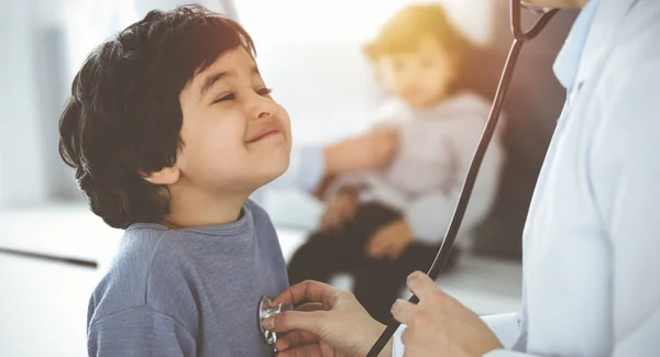 Médico-mujer examinando a un niño paciente por estetoscopio en Sunny Clinik. Lindo chico árabe y su hermano en la cita médica — Foto de Stock