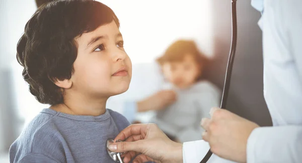 Ärztin untersucht eine Kinderpatientin per Stethoskop in einer sonnigen Klinik. Niedlicher arabischer Junge und sein Bruder beim Arzttermin — Stockfoto