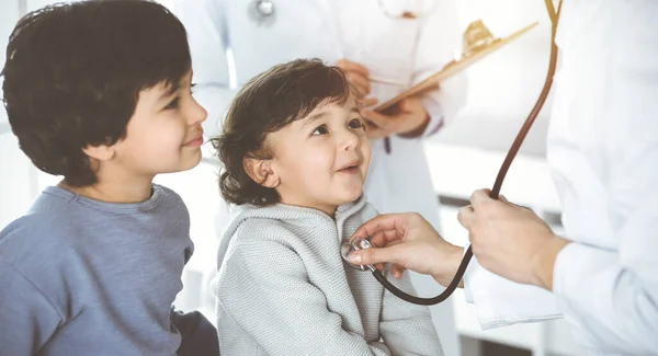 日当たりの良いクリニックで聴診器で小児患者を検査医師-女性.かわいいアラブ少年と彼の弟に予約時に医師 — ストック写真