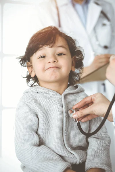 Mulher-médico examinando uma criança paciente por estetoscópio em clinik ensolarado. Criança árabe bonito na consulta médica — Fotografia de Stock