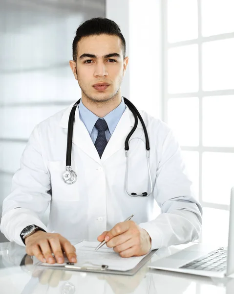 Arabo medico uomo sta utilizzando il computer portatile durante la compilazione di record di farmaci modulo presso la scrivania di vetro in r clinica. Concetto di medicina — Foto Stock