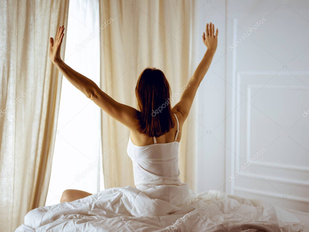 Woman stretching in bed after waking up, back view, entering a day happy and relaxed after good night sleep. Good morning and new day for brunette. Concept of Weekend, holidays