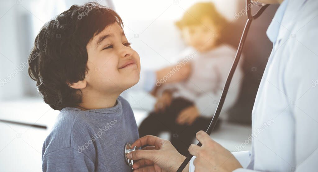 Doctor-woman examining a child patient by stethoscope in sunny clinik. Cute arab boy and his brother at physician appointment