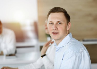 Neşeli gülümseyen iş adamı güneşli ofiste çalışıyor. Sıradan giyimli girişimci arka planda çeşitli meslektaşlarıyla otururken bilgisayar kullanıyor