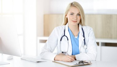 Güneşli klinikte masa başında oturan kadın doktor. Sarışın, neşeli doktor ilaç geçmişi formunu dolduruyor.