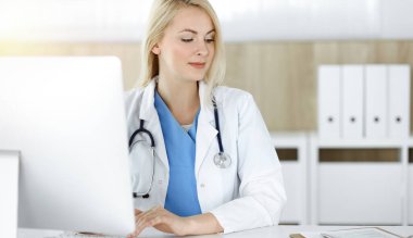 Güneşli klinikte masa başında oturan kadın doktor. Masaüstü bilgisayarı kullanan sarışın neşeli doktor