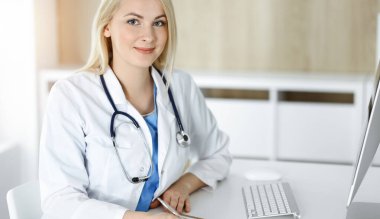 Güneşli klinikte masa başında oturan kadın doktor. Sarışın, neşeli doktor ilaç geçmişi formunu dolduruyor.
