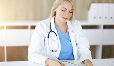 Güneşli klinikte masa başında oturan kadın doktor. Sarışın, neşeli doktor ilaç geçmişi formunu dolduruyor.