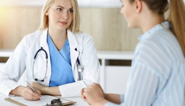 Kadın doktor ve hasta güneşli klinikte oturup konuşuyor. Sarışın terapist neşeyle gülümsüyor.