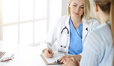 Kadın doktor ve hasta güneşli klinikte oturup konuşuyor. Sarışın terapist neşeyle gülümsüyor.